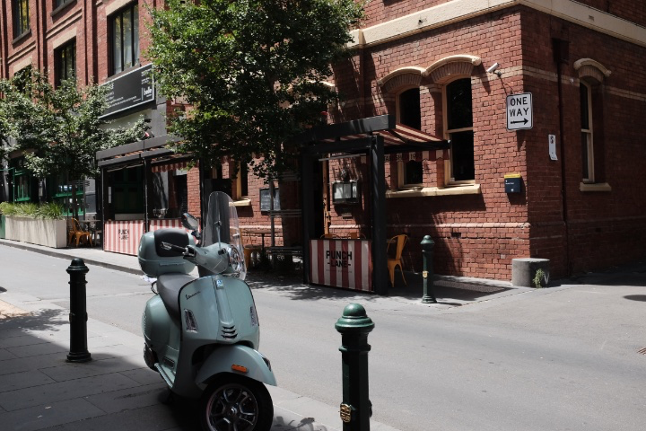 A scooter sitting in the shade while a brick building is across the street behind it.